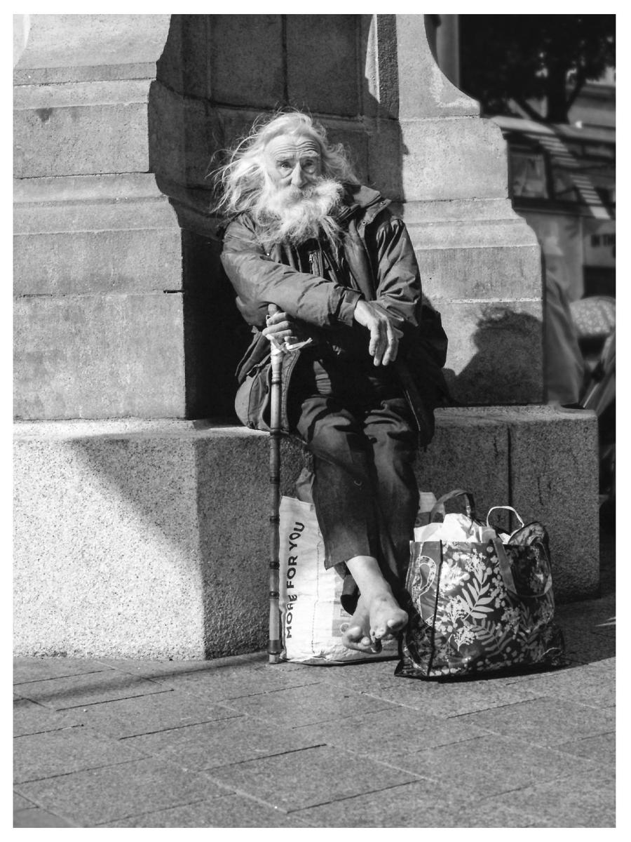 A man with long hair and a beard sits barefoot on a stone ledge with a walking stick and bags beside him.