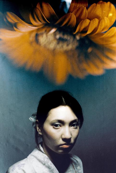 A woman with dark hair looks directly at the viewer, with a large orange flower above her head.