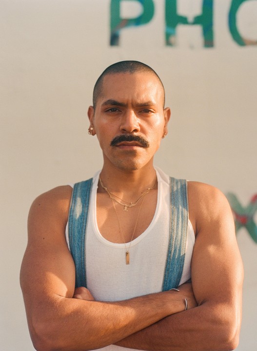 A man with a mustache and shaved head wears a white tank top and denim overalls with his arms crossed.