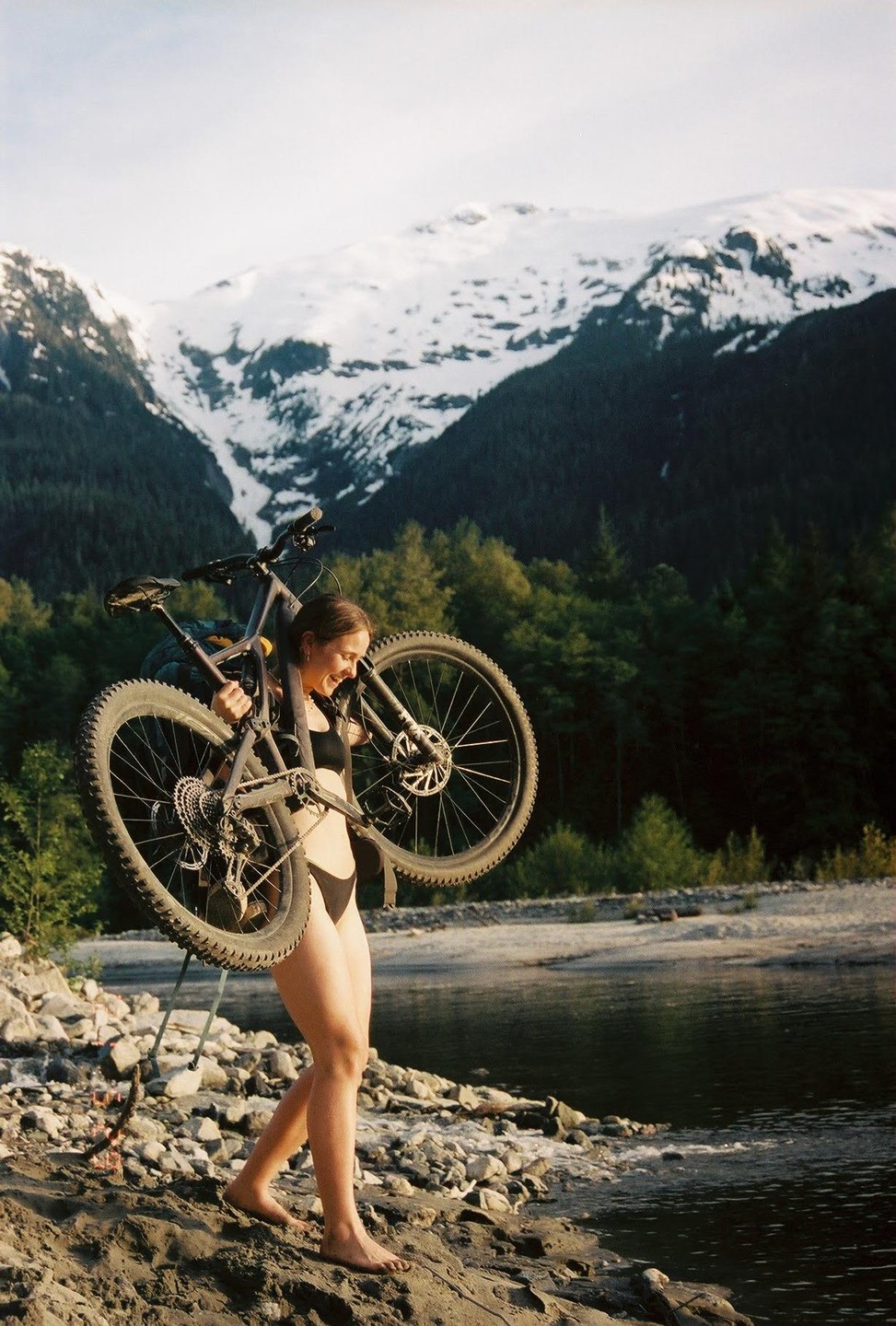 A woman carries a bicycle on her shoulder while walking barefoot along a rocky riverbank with snow-capped mountains and a forest in the background.