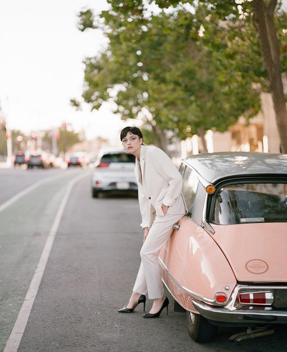 A woman in a white suit leans against a pink vintage car on a street.