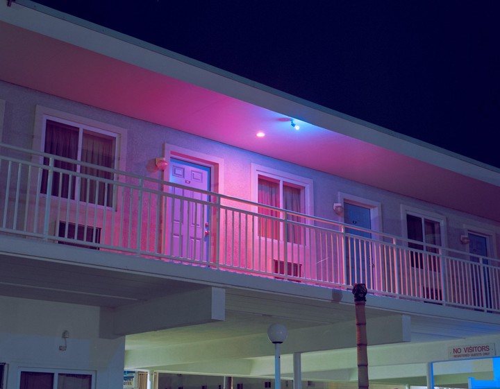 A motel balcony is illuminated by pink and blue neon lights at night.