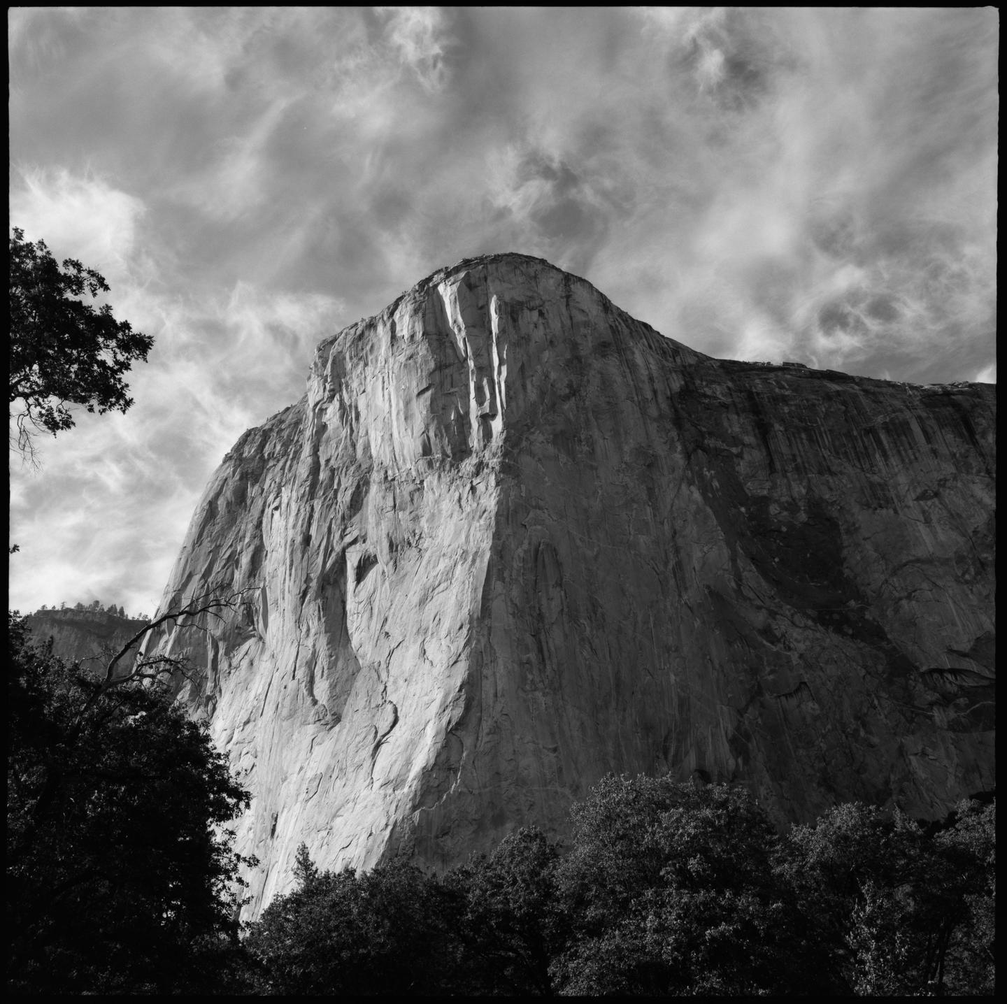 A large rock formation rises into the sky, with trees in the foreground and clouds above.