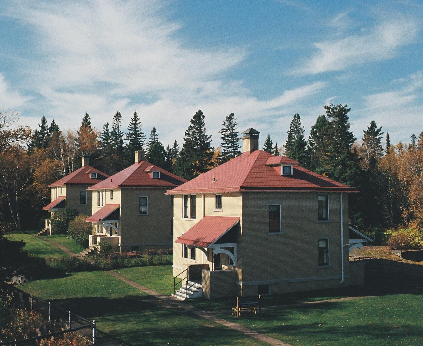 Three identical brick houses with red roofs are nestled among trees with autumn foliage under a blue sky.