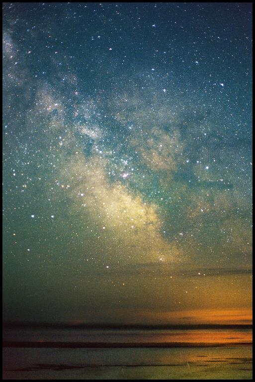 The milky way is visible above a body of water with a bright orange glow on the horizon.
