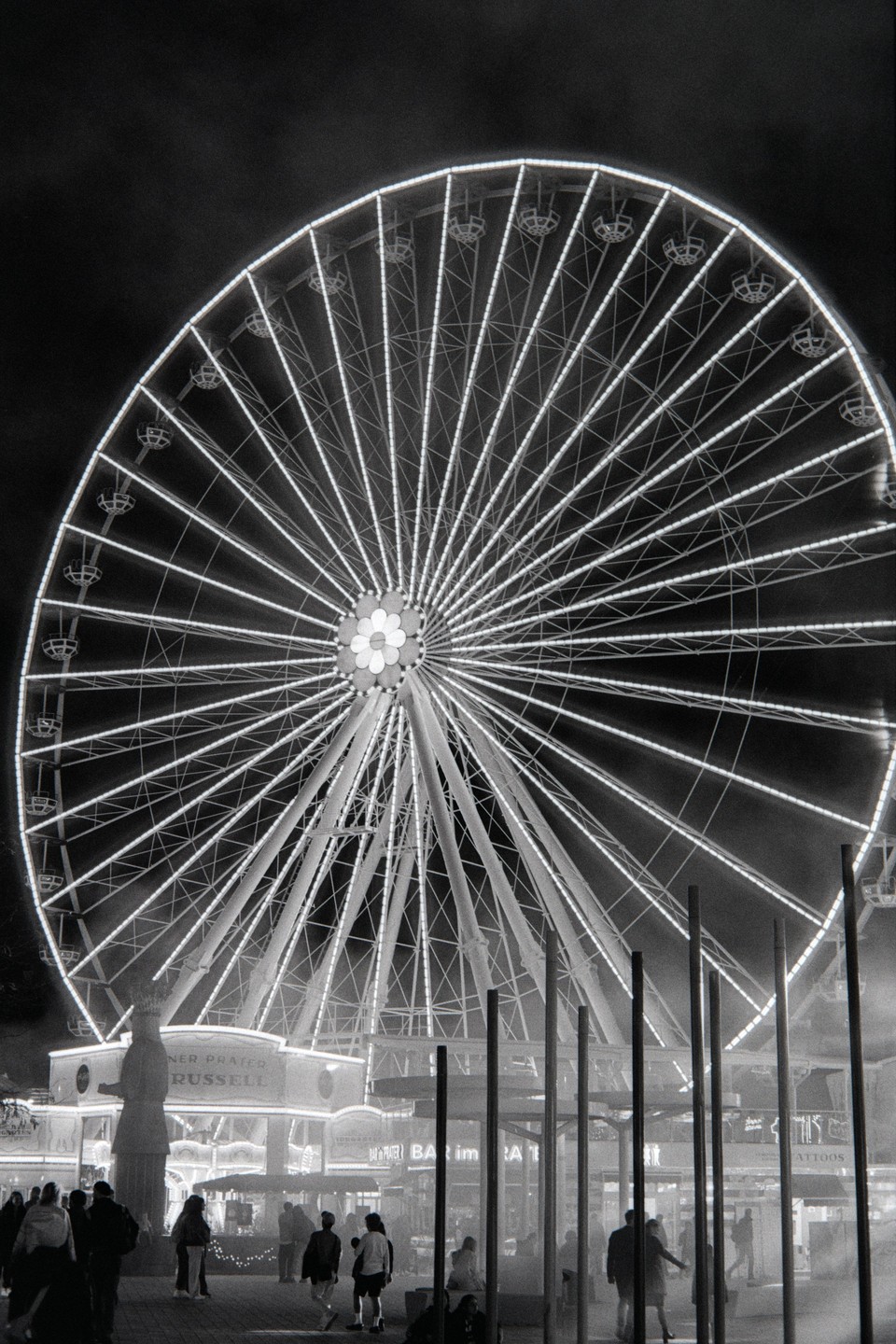 A large Ferris wheel illuminated at night towers over people walking in an amusement park.