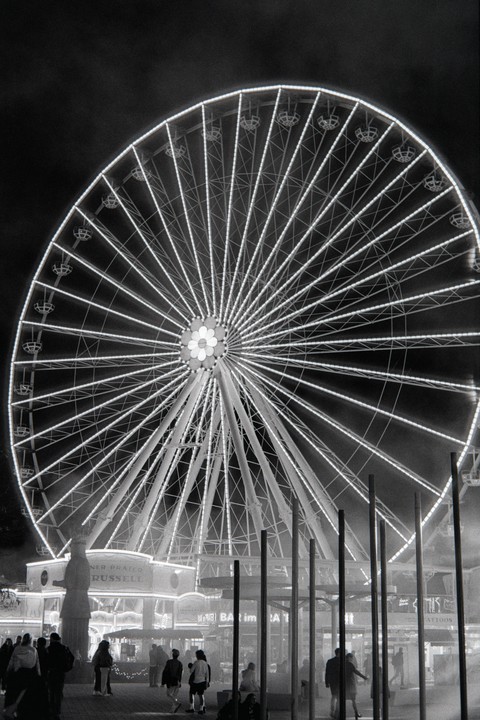 A large Ferris wheel illuminated at night towers over people walking in an amusement park.