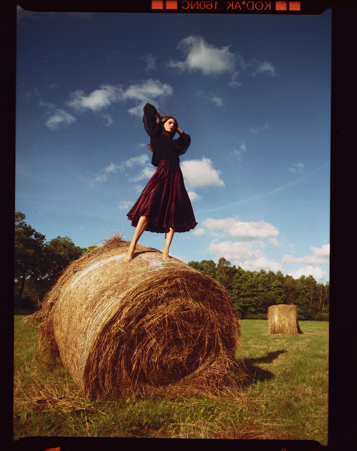 A woman stands barefoot on a large hay bale in a field under a blue sky with clouds.