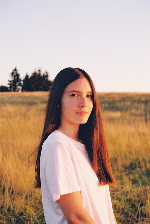 A young woman with long dark hair stands in a field of tall grass during golden hour.