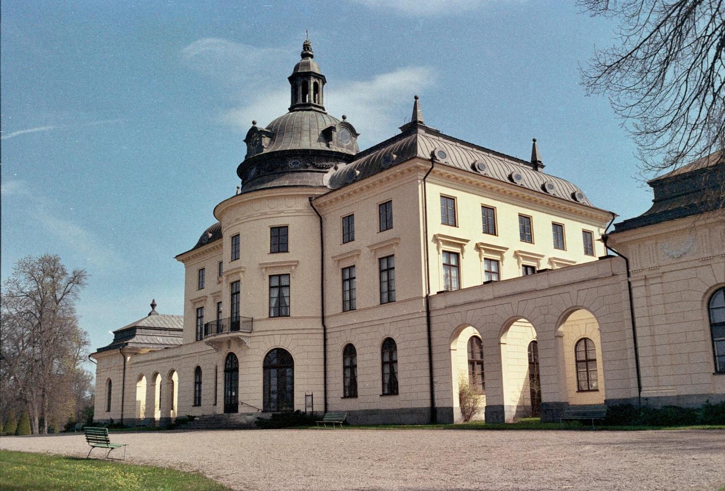 A large, ornate building with a domed roof and arched windows stands under a clear sky, with a gravel path and green grass in the foreground.