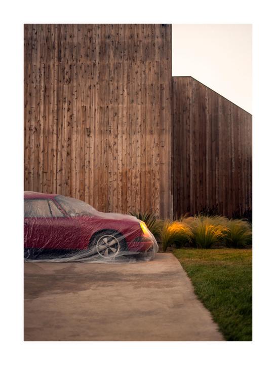 A red car covered in plastic is parked on a concrete driveway next to a wooden building.