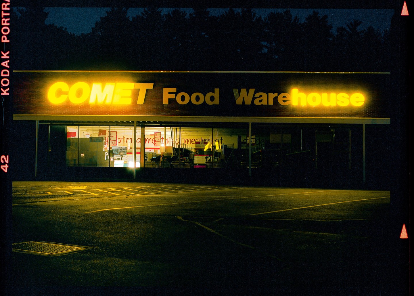 A Comet Food Warehouse store is illuminated by its neon sign at night, with a parking lot in the foreground.