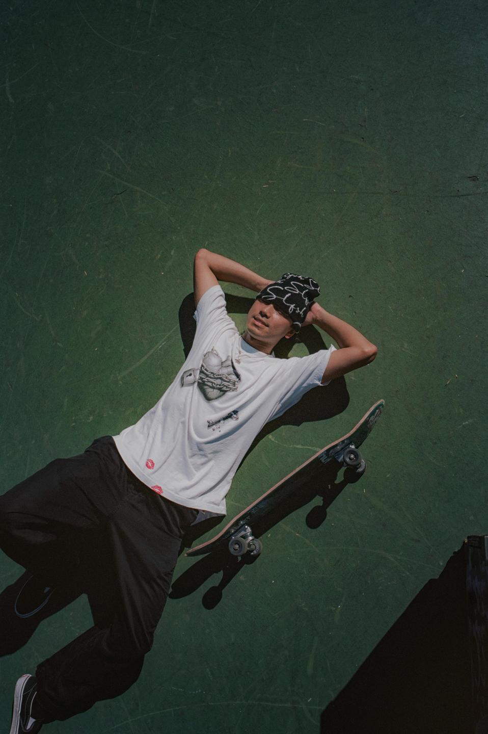 A young man wearing a beanie and t-shirt lies on a green surface with a skateboard next to him.