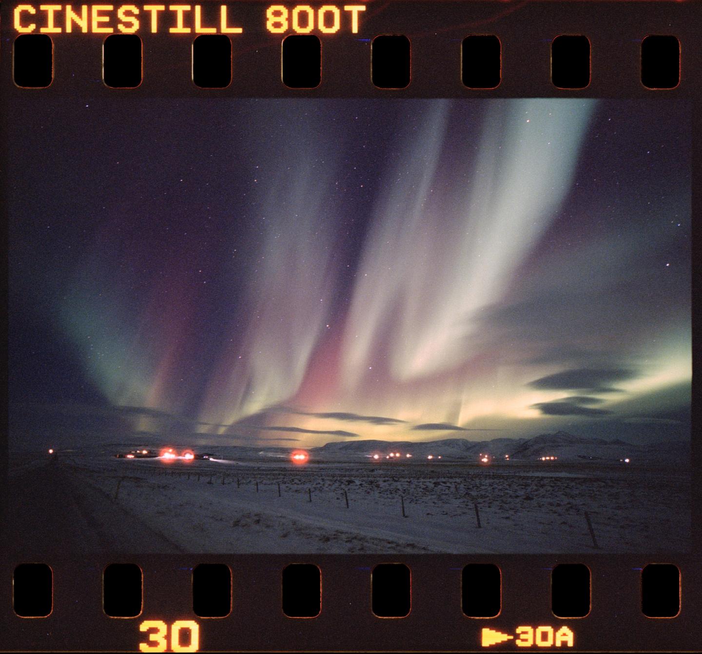 The aurora borealis illuminates a snowy landscape with distant lights under a starry night sky.