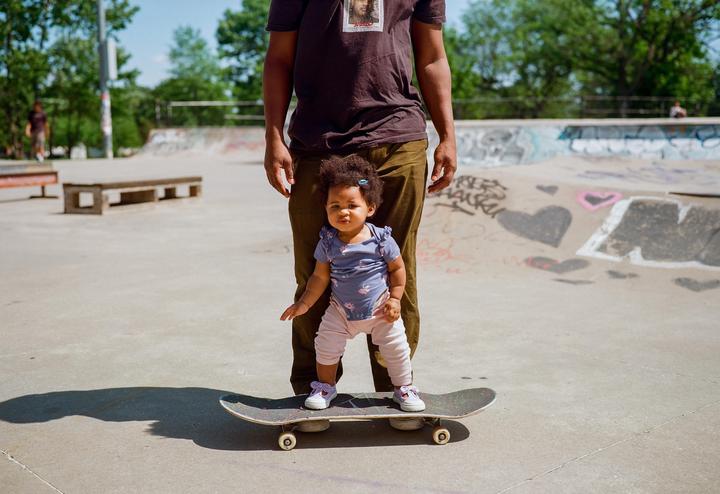 A baby stands on a skateboard in a skatepark with an adult standing behind them.