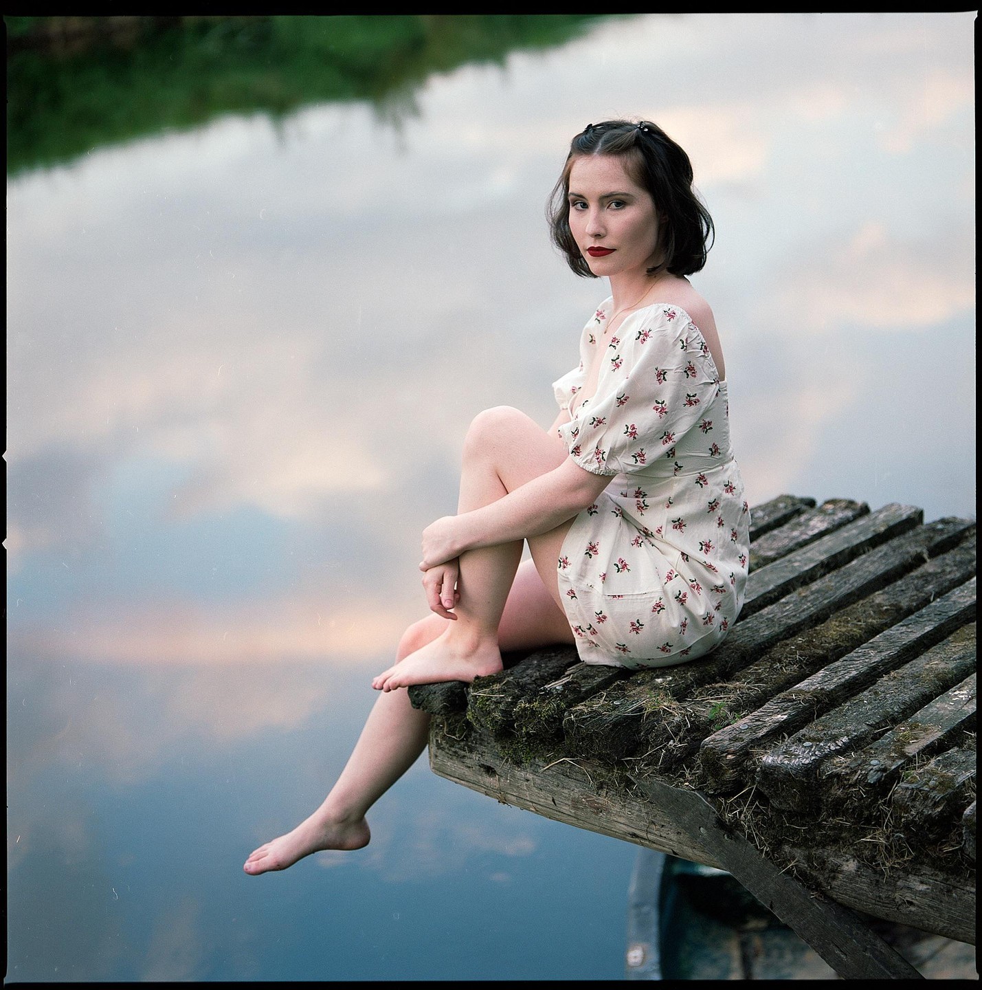 A woman in a floral dress sits on a wooden dock with her feet dangling over the water.