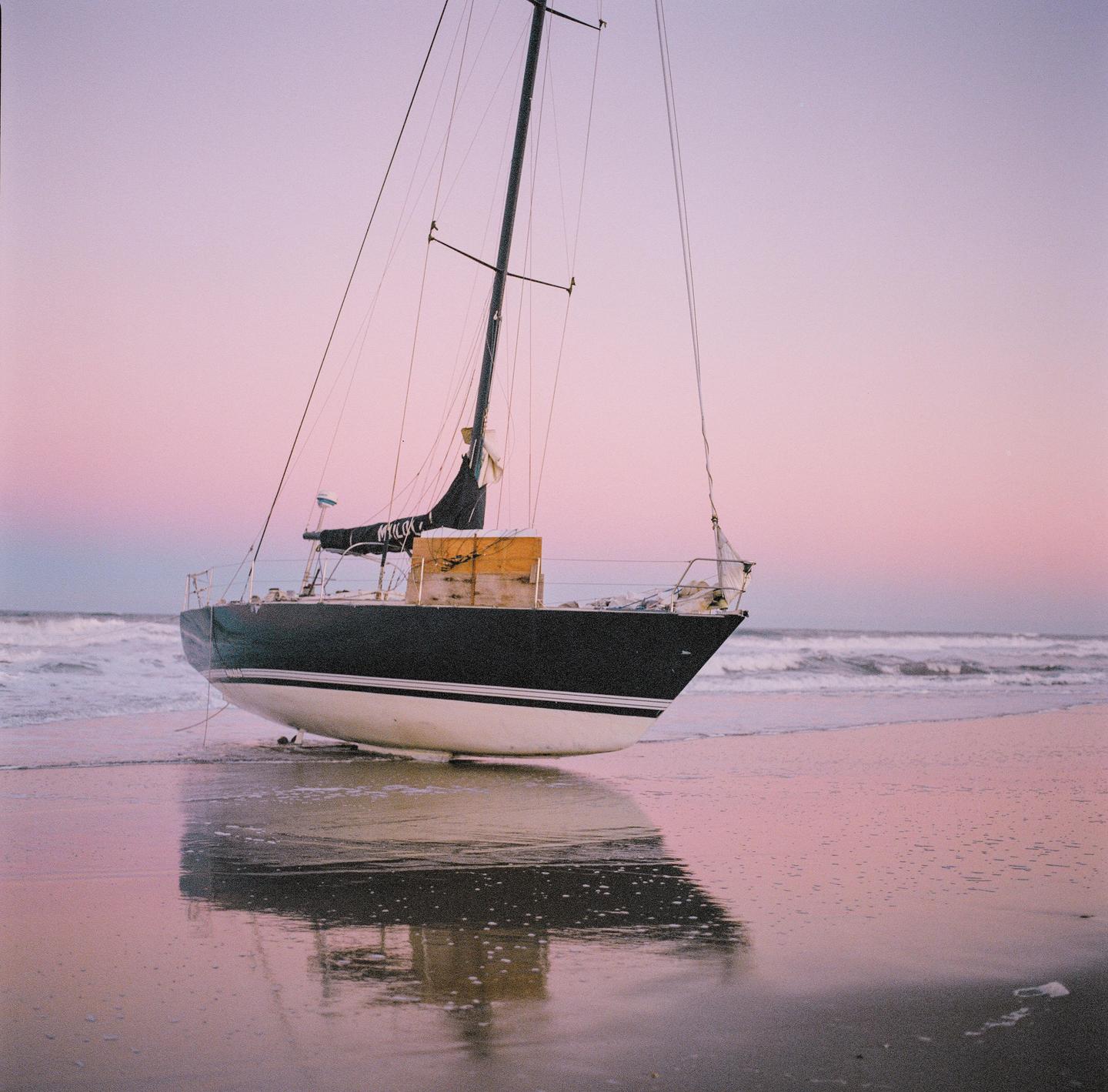 A sailboat is beached on the sand with waves in the background under a pink sky.