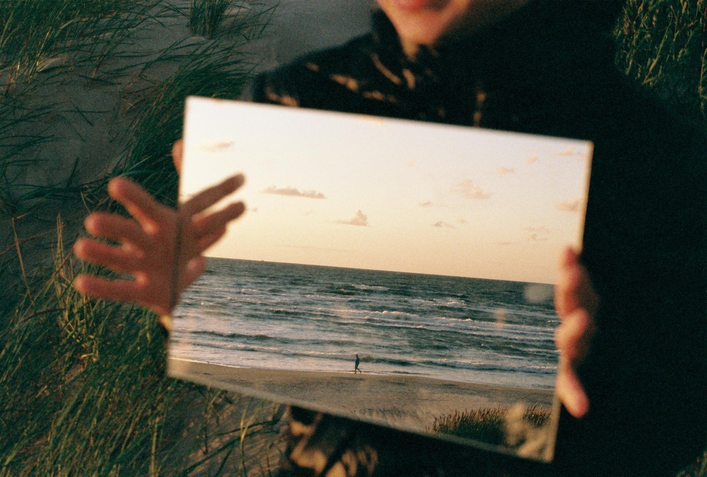 A person holds a mirror reflecting a beach scene with a lone figure walking by the water at sunset.
