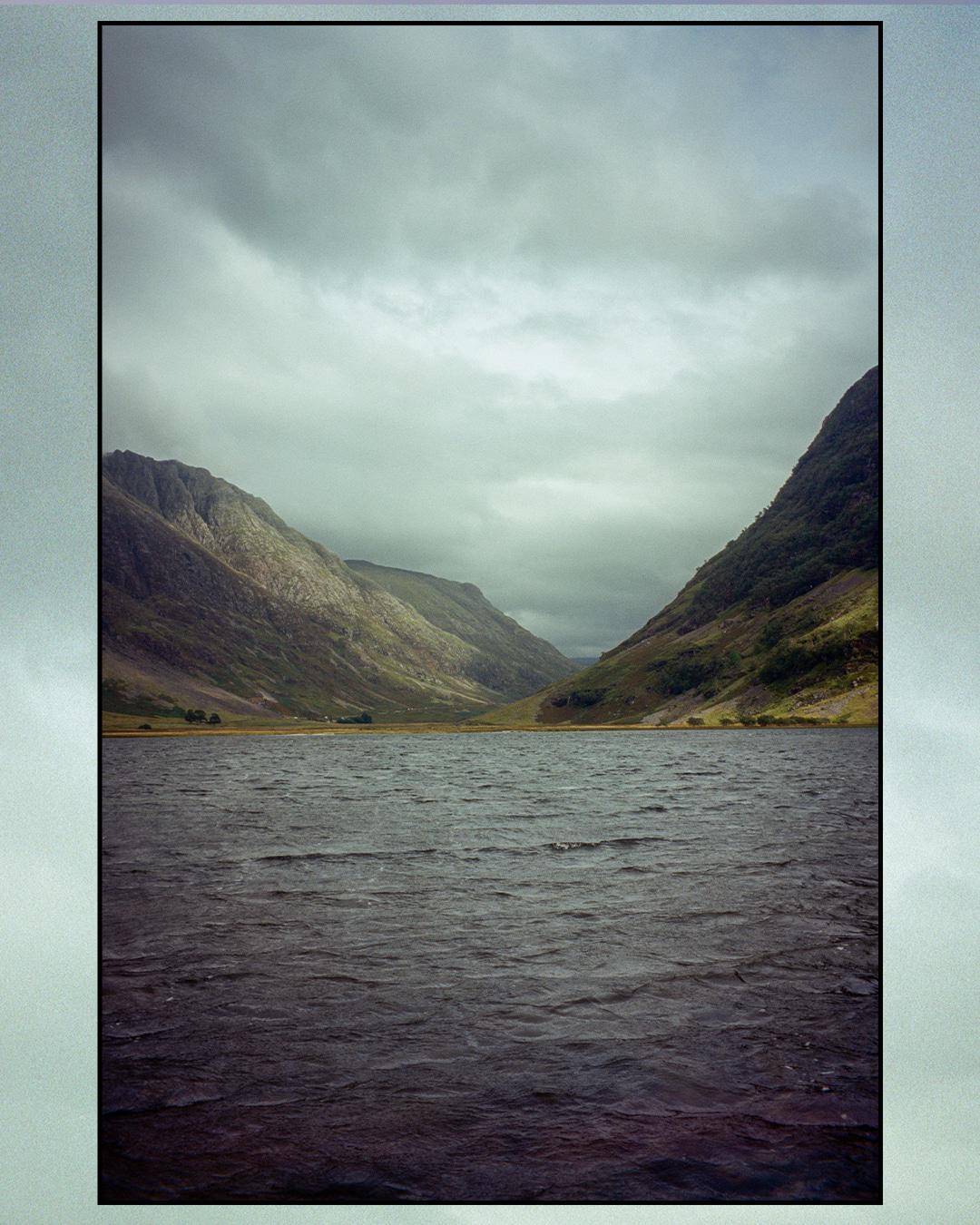 A body of water with ripples on its surface is surrounded by mountains under a cloudy sky.