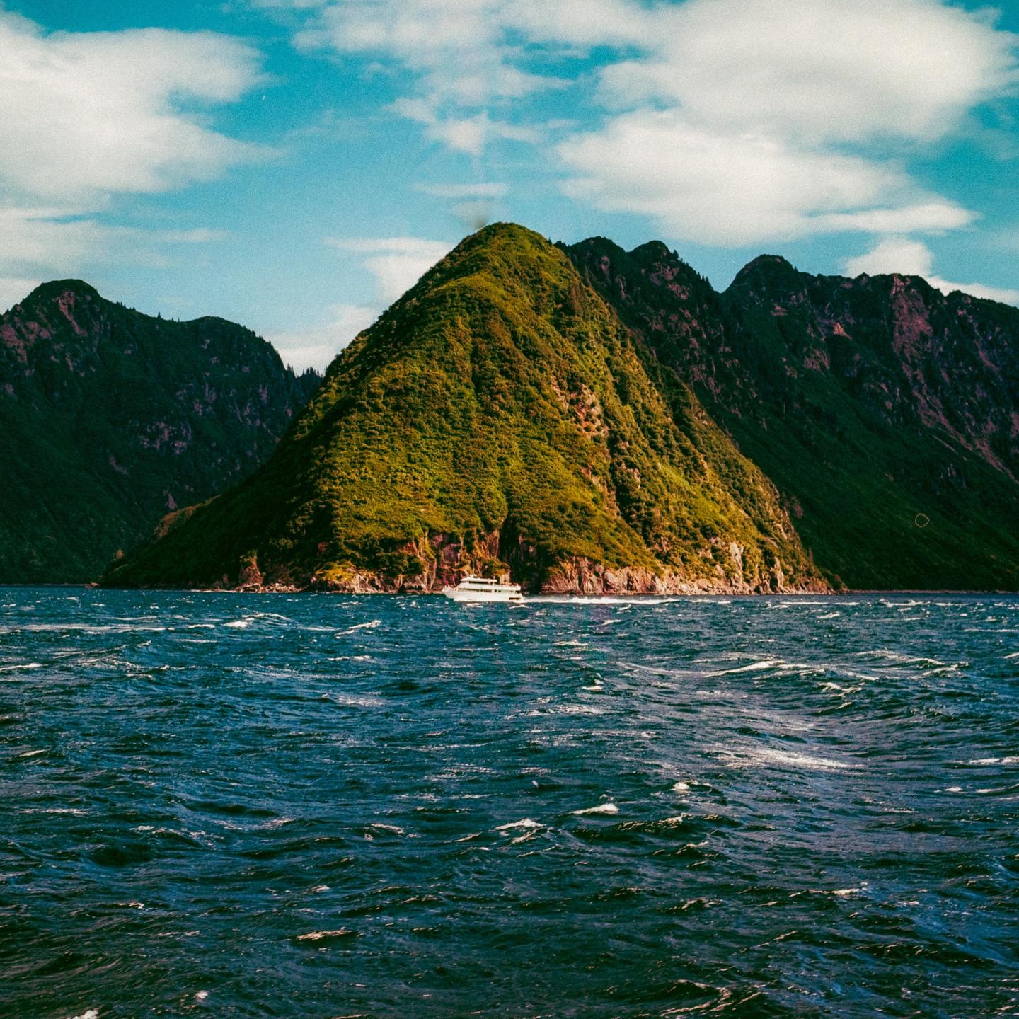 A white boat travels across choppy water with a large, green, conical mountain in the background under a blue sky with clouds.