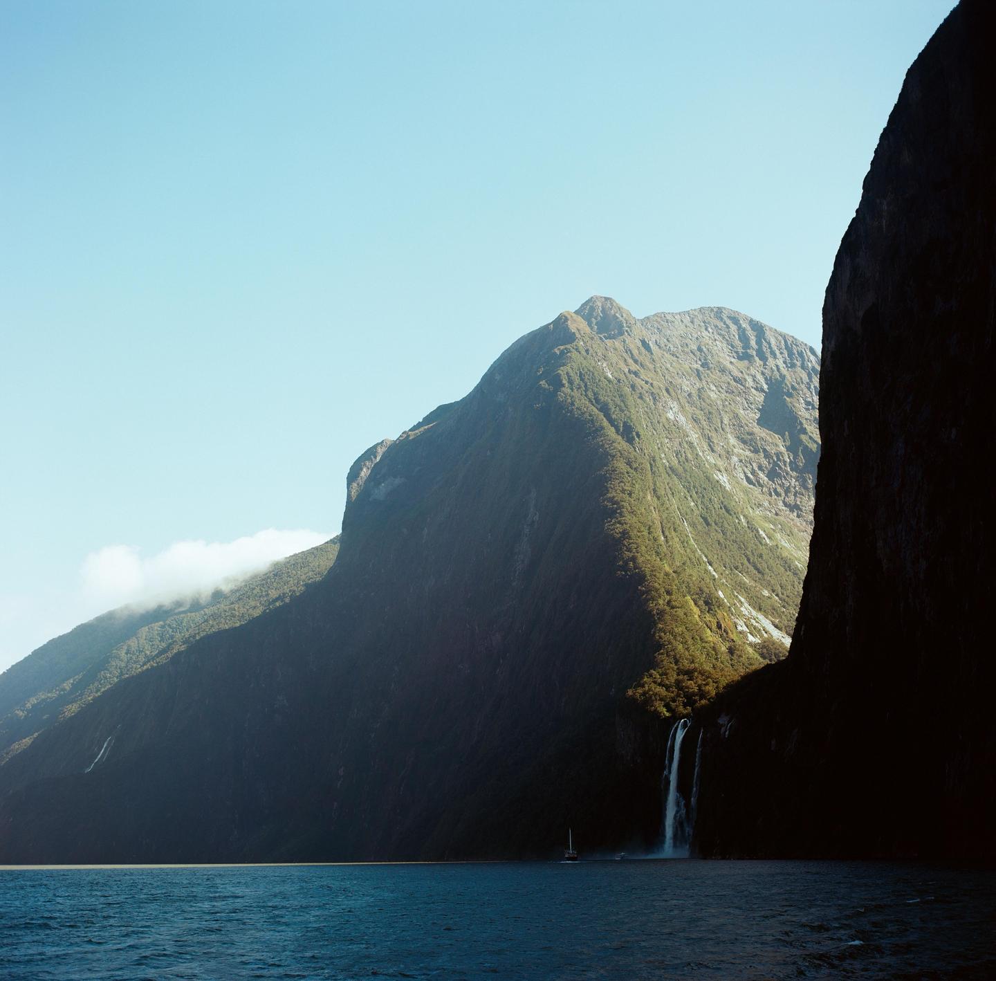 A waterfall cascades down a large mountain into a body of water with a small boat.