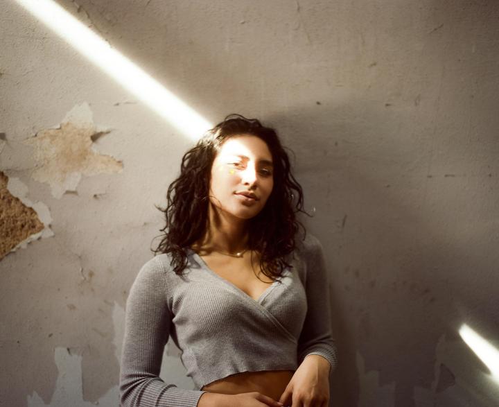 A woman with dark, curly hair and a gray top stands against a distressed wall with a beam of light across her face.