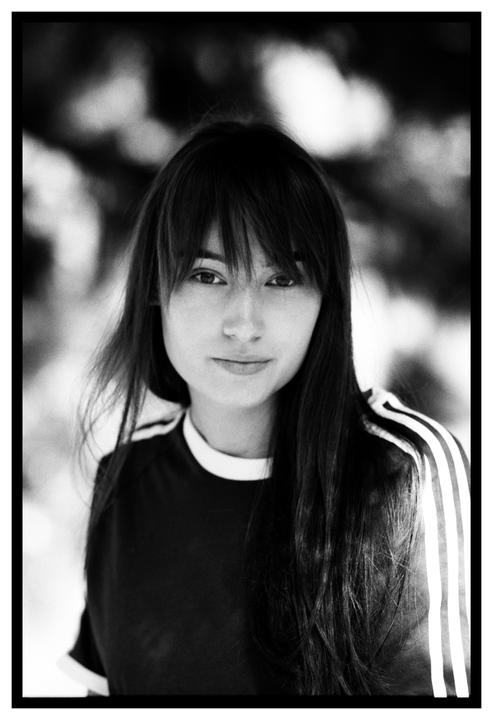 A woman with long hair and bangs looks directly at the viewer in a black and white portrait.