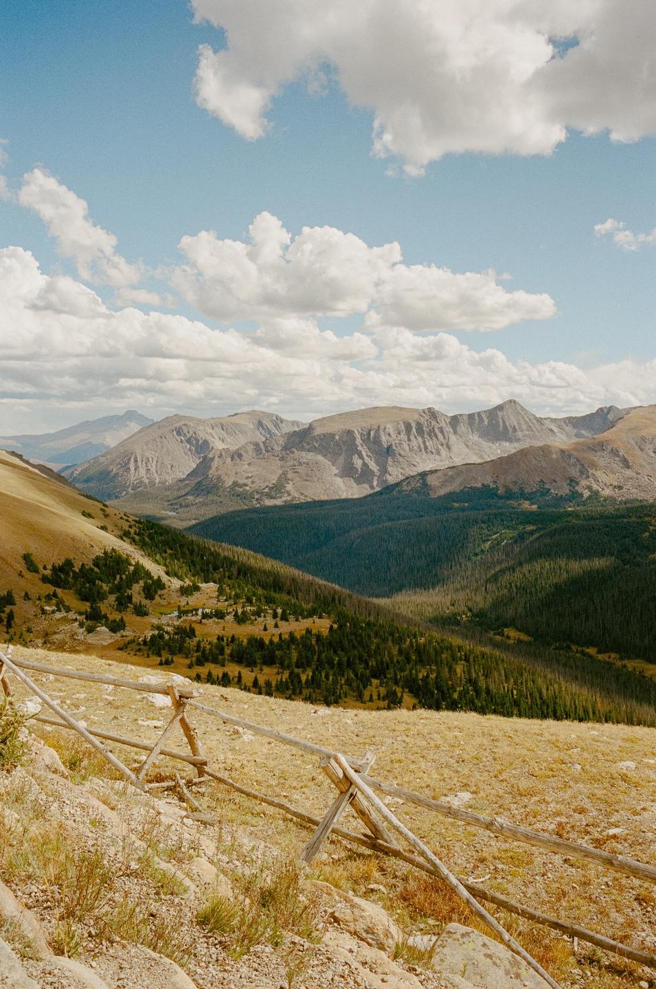 A wooden fence runs along a mountain slope with a valley and more mountains in the distance under a cloudy sky.