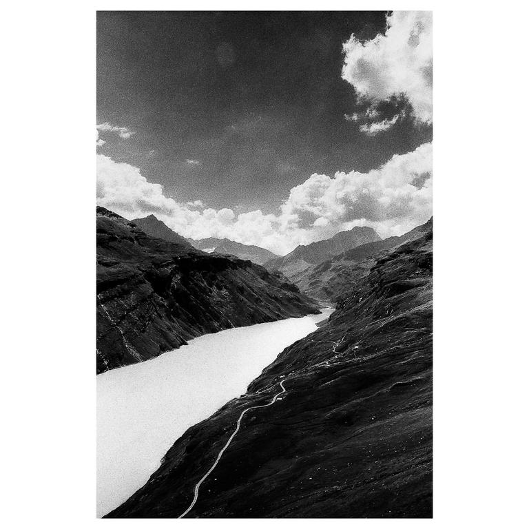 A winding road follows a body of water through a mountainous landscape under a cloudy sky.