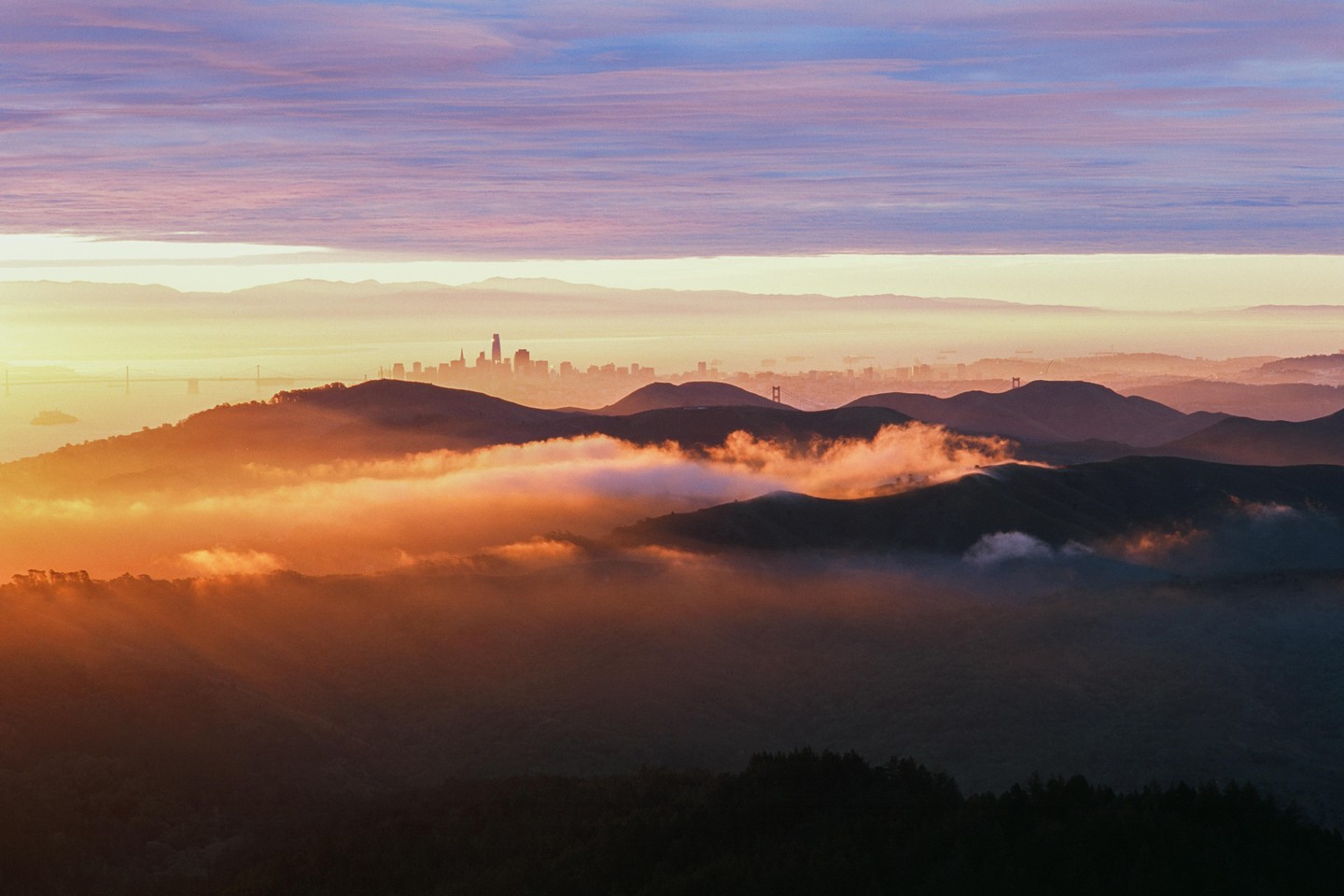 A sunrise over a mountain range with a city skyline and bridges visible in the distance, with fog filling the valleys.