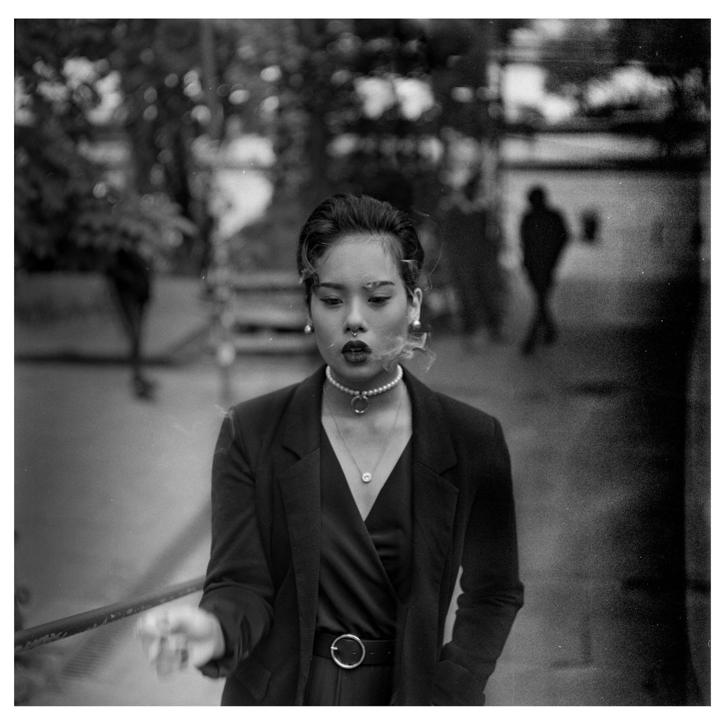 A woman with a pearl necklace and a dark blazer smokes a cigarette, with smoke coming from her mouth.