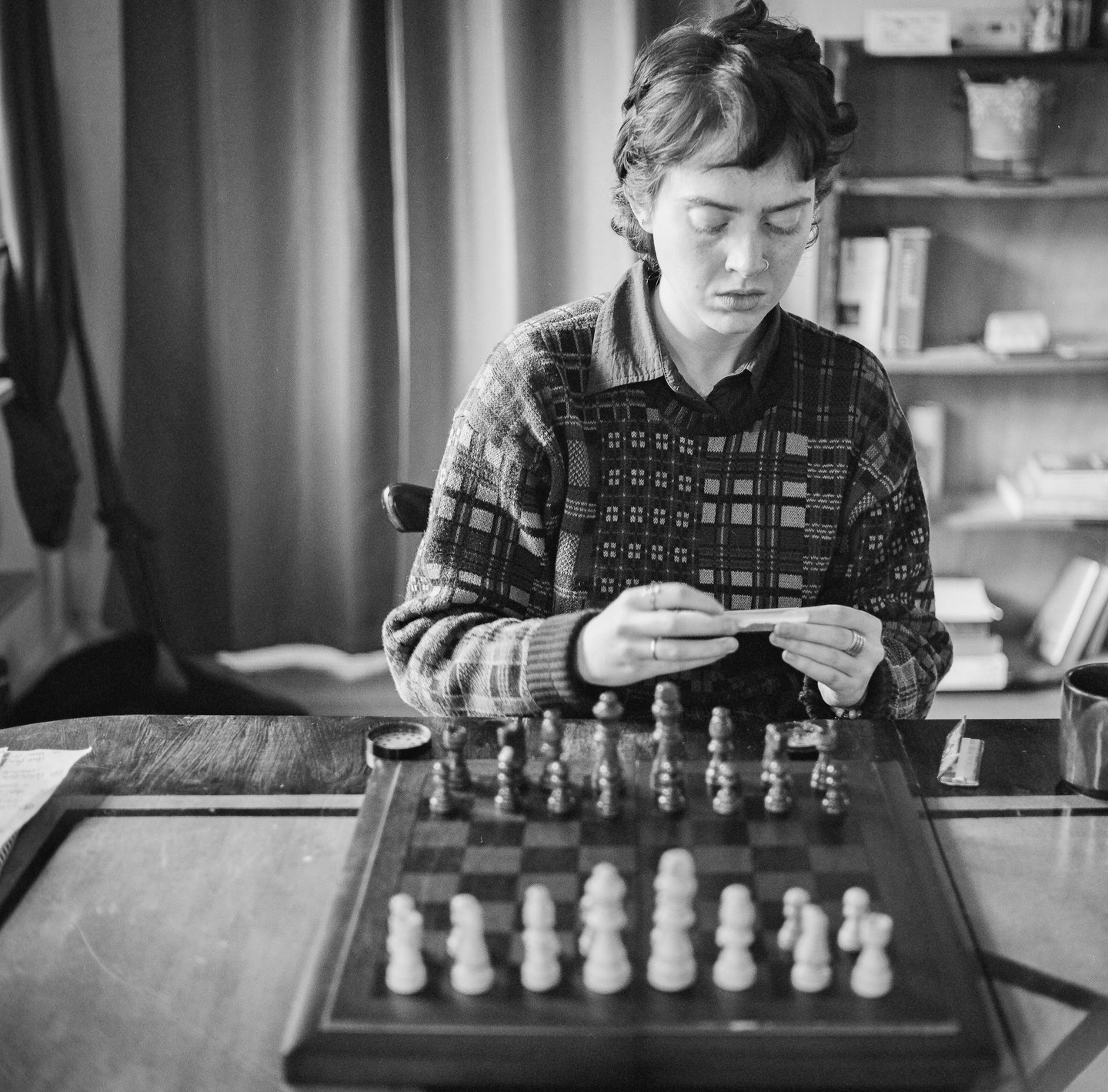 A woman sits at a table with a chessboard, rolling a joint.