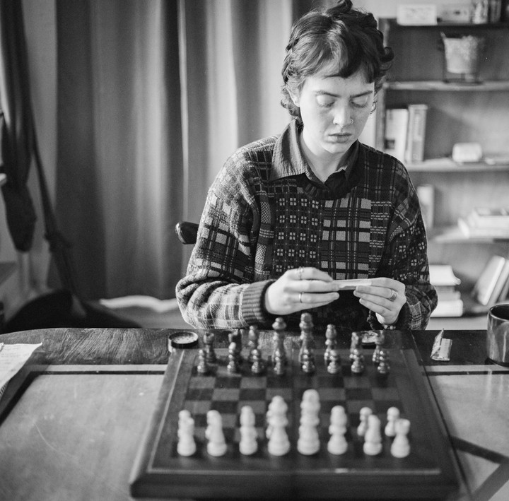 A woman sits at a table with a chessboard, rolling a joint.