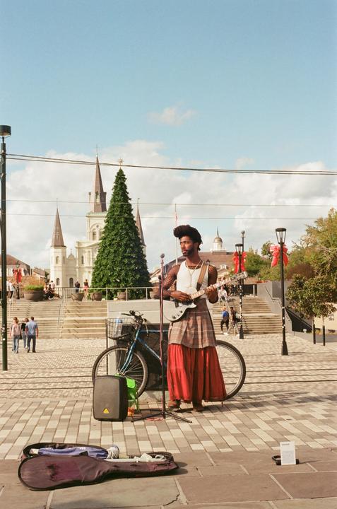 A man plays guitar on a paved street with a church and Christmas tree in the background.