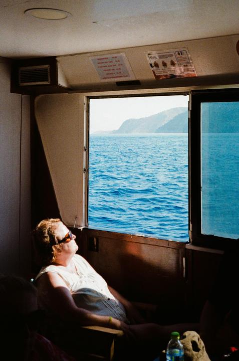 A woman wearing sunglasses sits in a boat looking out an open window at the ocean and distant mountains.