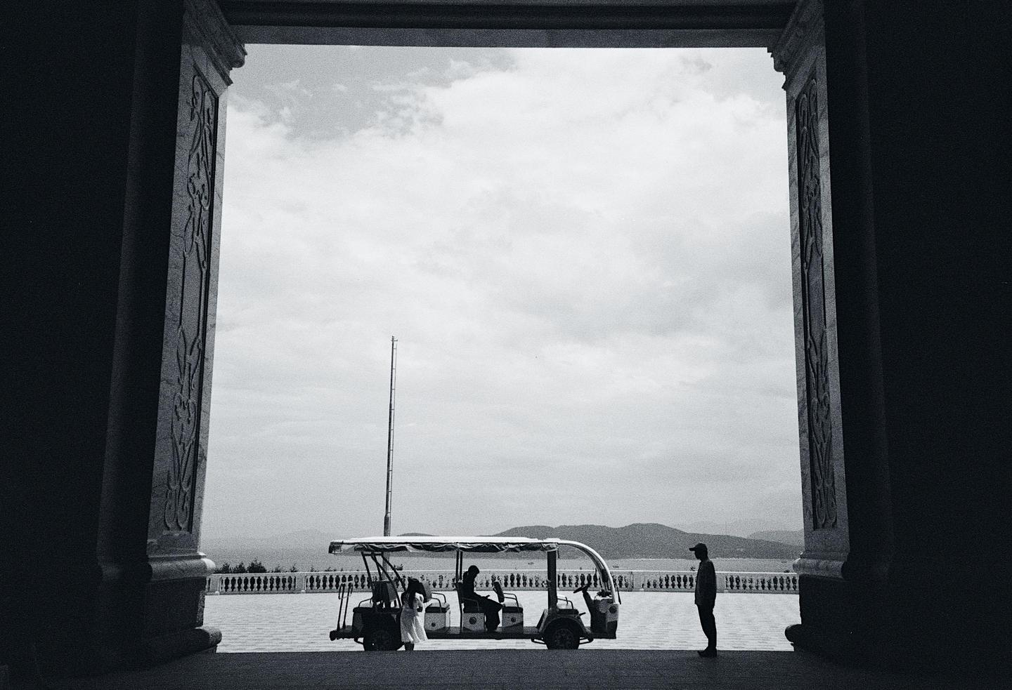 A golf cart with people is framed by a dark archway overlooking a body of water and mountains.