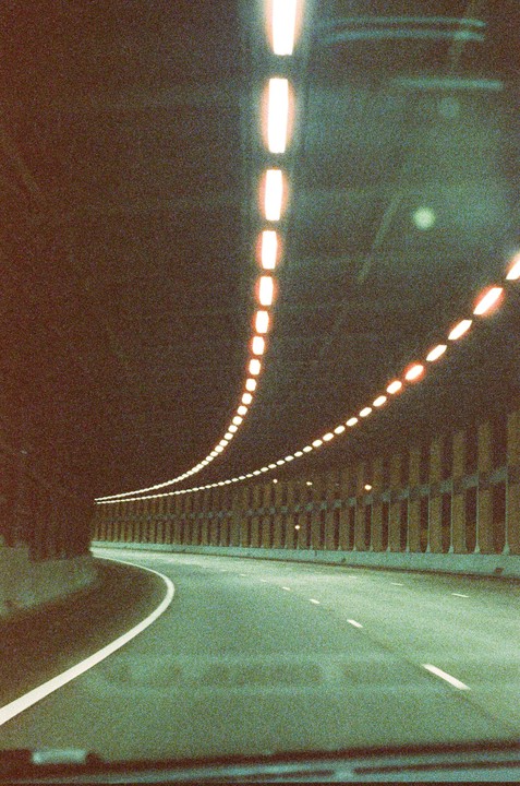 A long exposure shot from inside a vehicle shows a winding road through a tunnel at night, with a line of lights along the ceiling.