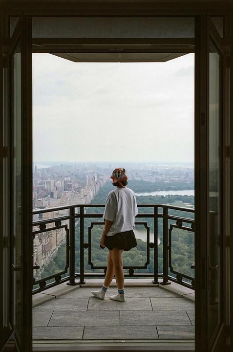 A woman stands on a balcony overlooking a city park and skyline.