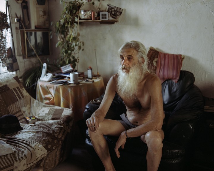 A man with a long white beard sits shirtless on a black armchair in a cluttered room.