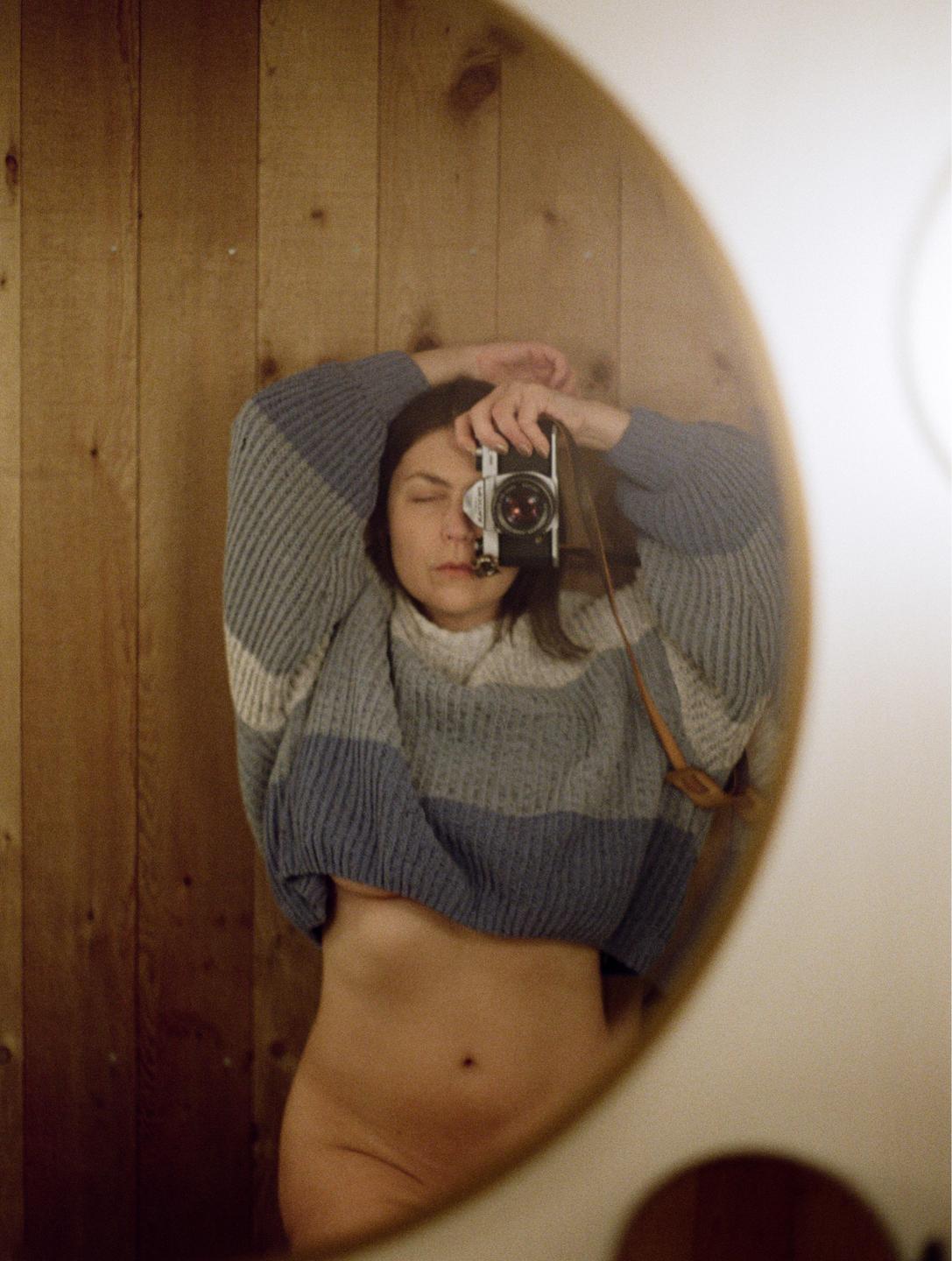 A woman in a striped sweater takes a mirror self-portrait with a camera, revealing her bare midriff and hip.