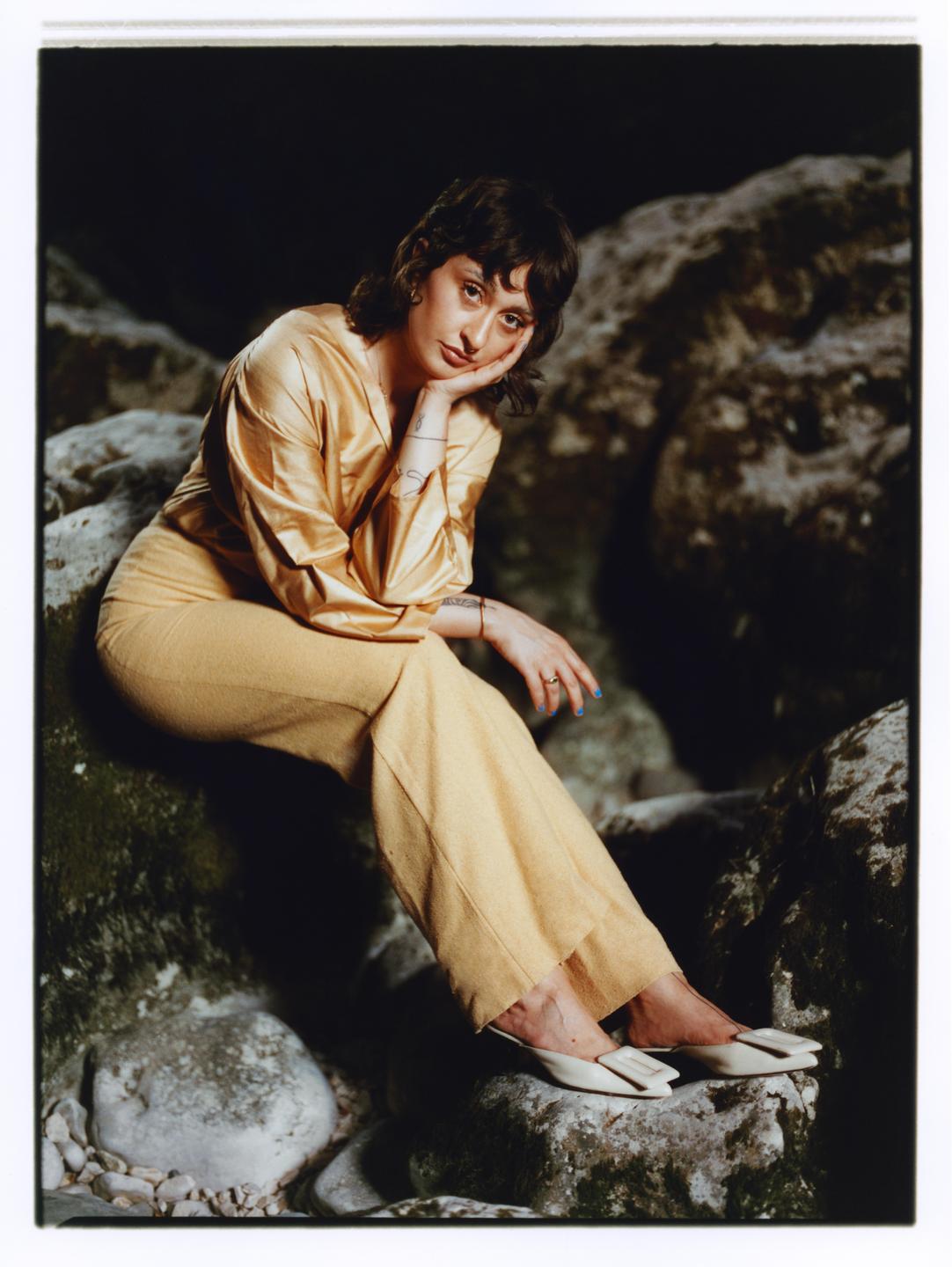 A woman in a gold shirt and pants sits on a rock.