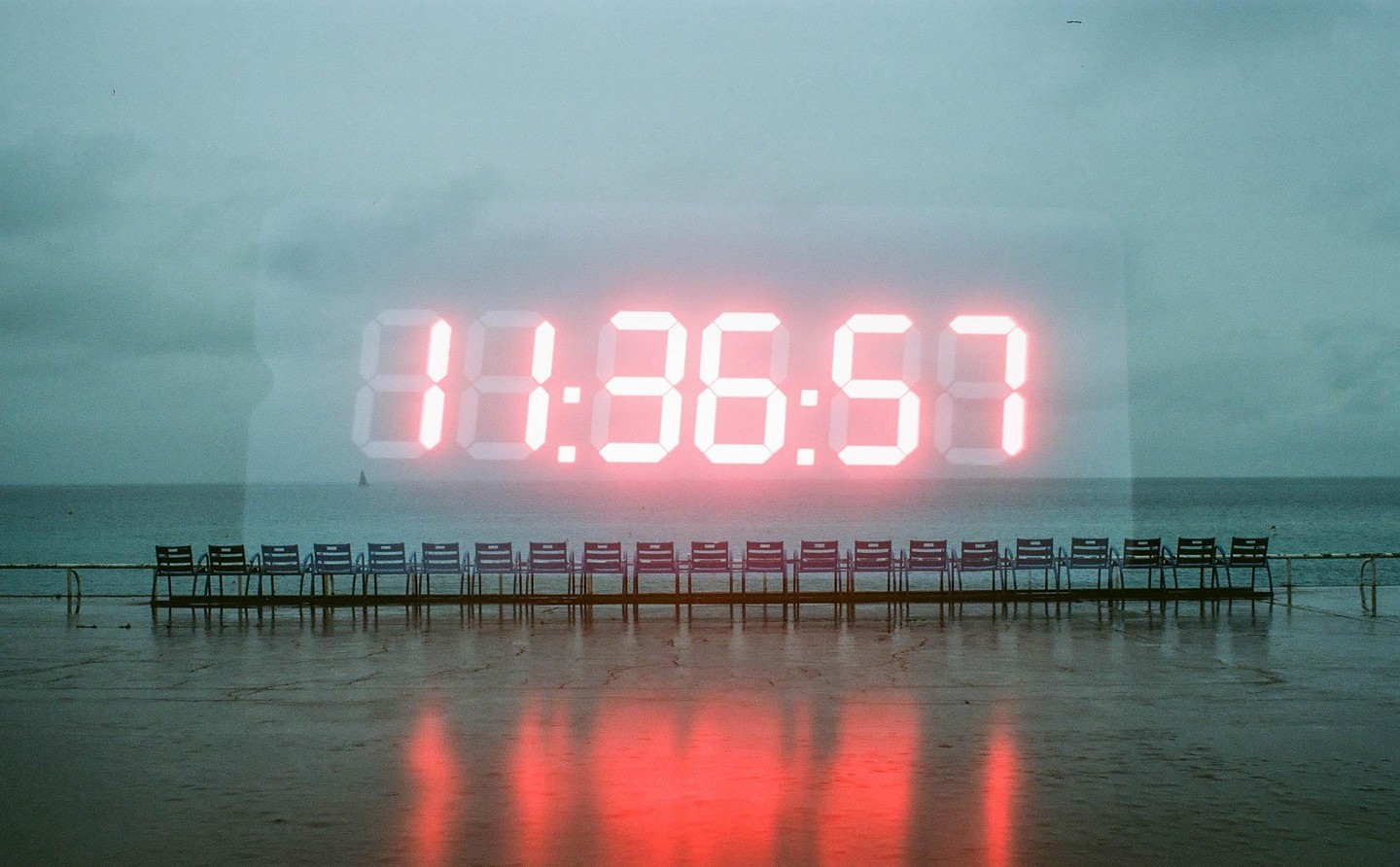 A digital clock showing 11:36:57 is double exposed over a row of chairs facing the ocean under a cloudy sky.