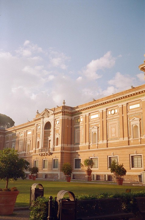A grand, ornate building with a large arched entrance and numerous windows overlooks a green lawn with potted trees and benches under a partly cloudy sky.