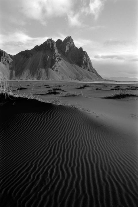 A black and white landscape shows a mountain range behind a sandy beach with ripples in the foreground.