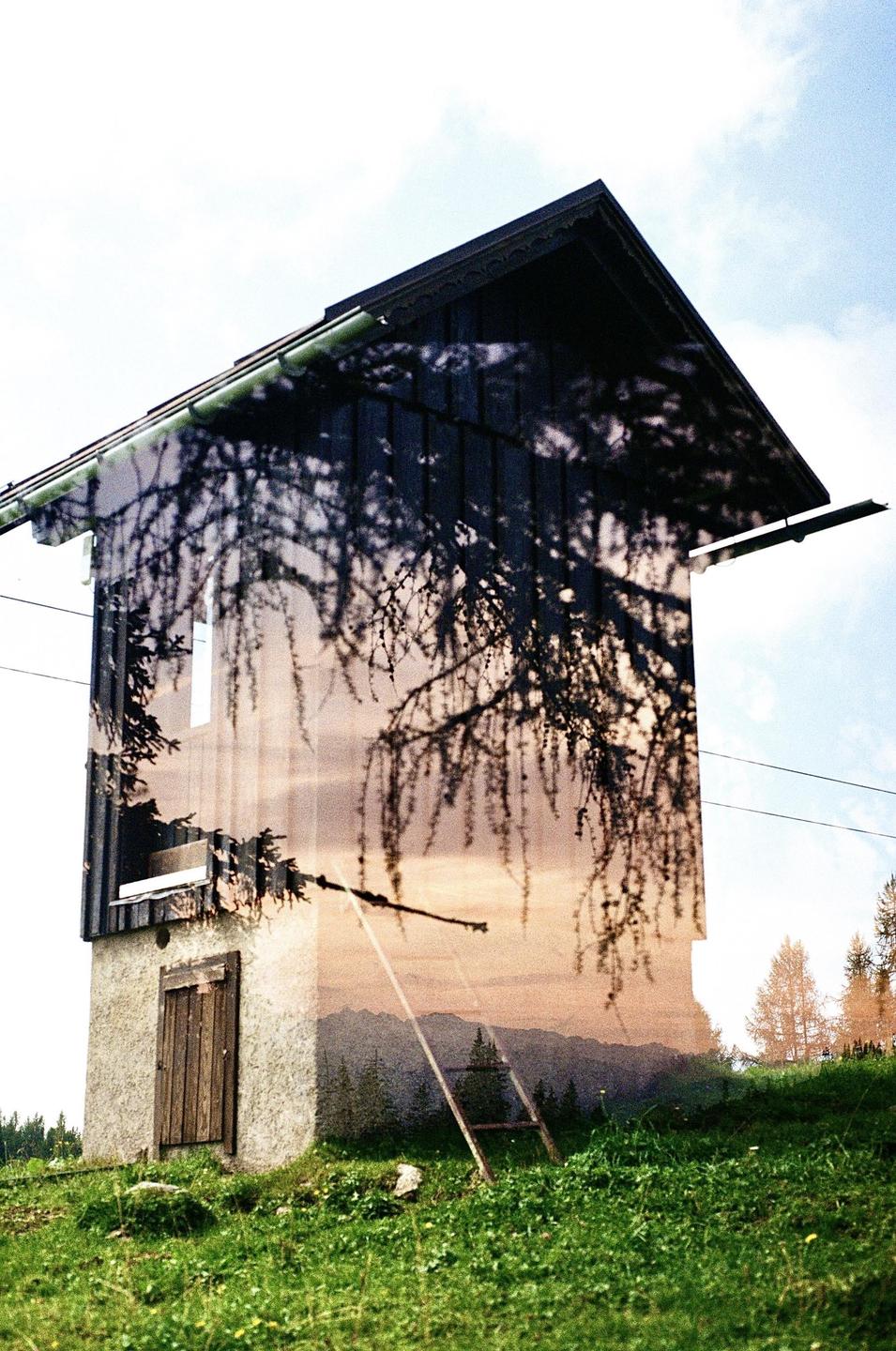 A double exposure shows a small building with a wooden door and a ladder leaning against it, superimposed with a mountain landscape and tree branches.