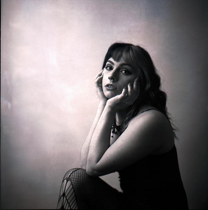 A woman with dark hair and a nose ring poses with her hands cupping her face.