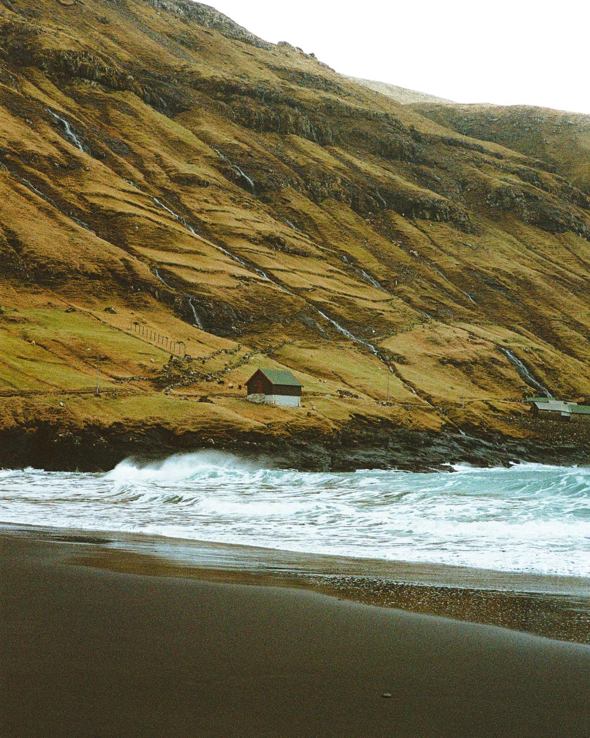 A small house with a green roof sits on a grassy hillside overlooking a black sand beach with crashing waves.