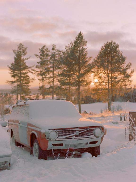 A snow-covered car is parked in a snowy landscape with pine trees and a sunset in the background.