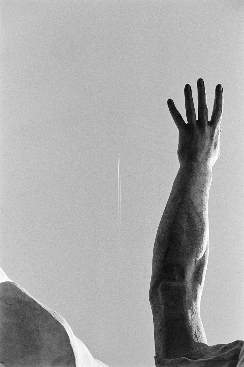 A statue's arm and hand reach towards the sky, with an airplane contrail visible in the distance.
