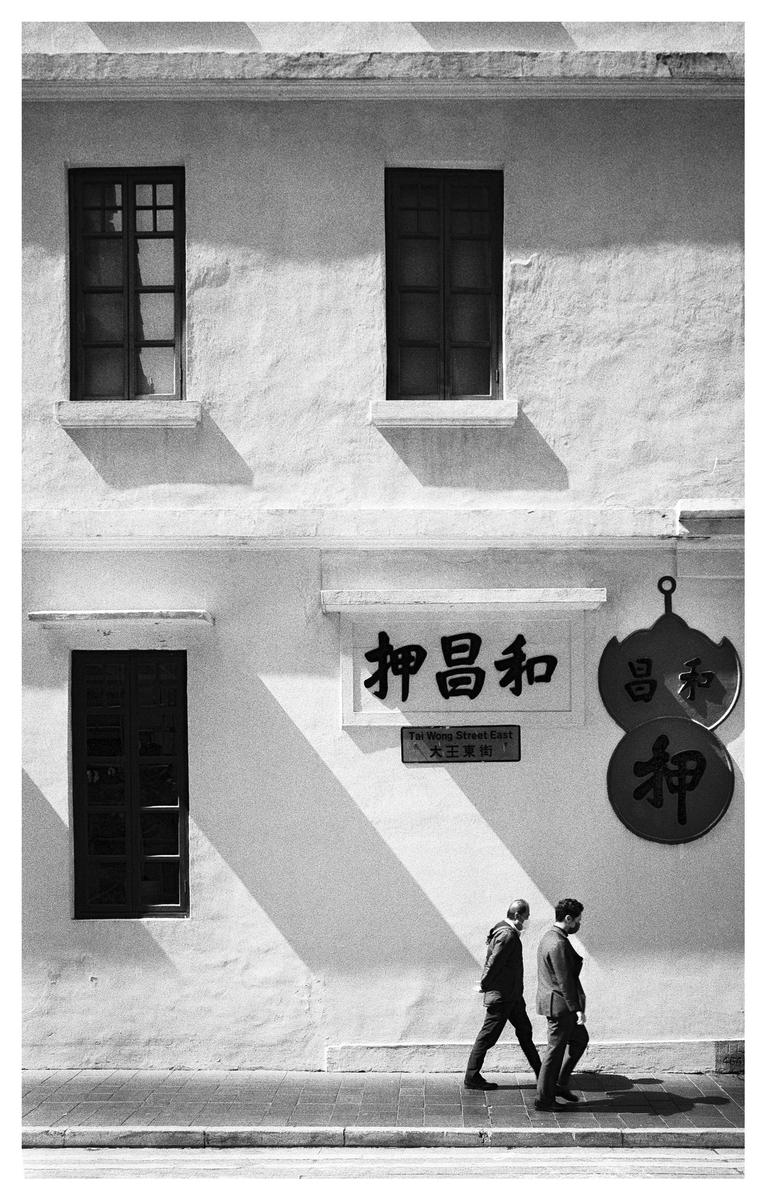 Two men walk on a sidewalk past a building with windows and signs in a street photography shot.